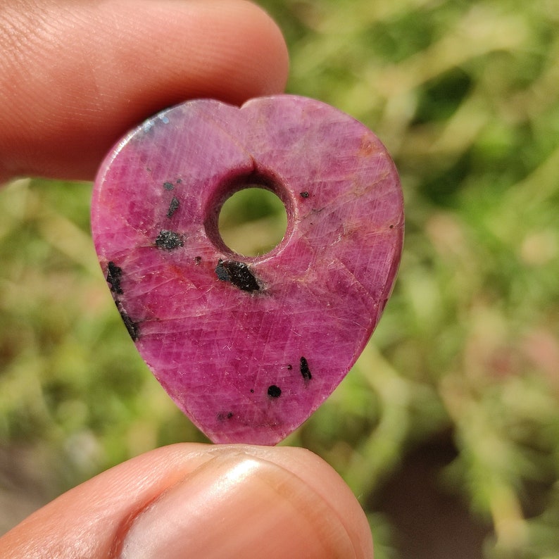 May include: A heart-shaped pink ruby pendant with a hole drilled through the top. The ruby has black inclusions throughout.