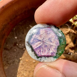 K&ouml;nnte beinhalten: Ein runder Edelstein mit einem zentralen lila Muster, umgeben von gr&uuml;nen und wei&szlig;en Schichten. Der Edelstein wird zwischen Fingern gehalten, was sein kompliziertes Design und seine nat&uuml;rlichen Farben hervorhebt. Es ist wahrscheinlich ein dekorativer Gegenstand.