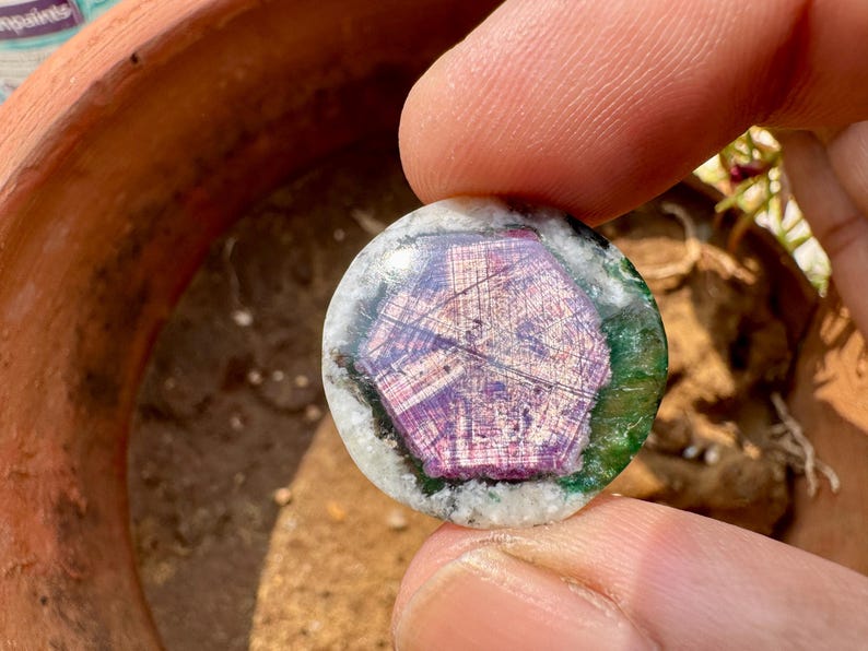 K&ouml;nnte beinhalten: Ein runder Edelstein mit einem zentralen lila Muster, umgeben von gr&uuml;nen und wei&szlig;en Schichten. Der Edelstein wird zwischen Fingern gehalten, was sein kompliziertes Design und seine nat&uuml;rlichen Farben hervorhebt. Es ist wahrscheinlich ein dekorativer Gegenstand.