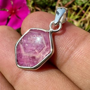 K&ouml;nnte beinhalten: Ein Anh&auml;nger mit einem sechseckigen, durchscheinenden rosa Edelstein mit sichtbaren geometrischen Innenmustern, gefasst in einen polierten silberfarbenen Metallrahmen mit einer kleinen runden &Ouml;se an der Oberseite.