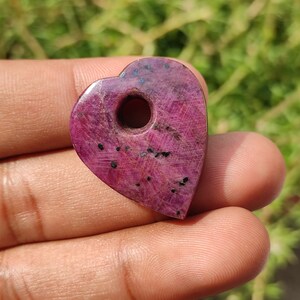 May include: A heart-shaped pendant made of pink ruby with a hole drilled through the top. The pendant has a rough, natural texture and is a deep pink color.