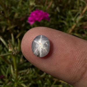 May include: A white star sapphire gemstone with a six-rayed star in the center. The gemstone is oval-shaped and has a smooth, polished surface.