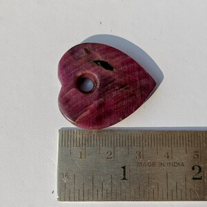 以下が含まれることがあります： 中央に穴が開いたハート型の木片。木は濃い紫色で、自然な木目模様があります。ハートは約3.8cm幅、5cm長さです。"Made in India"と書かれた定規の上に置かれています。