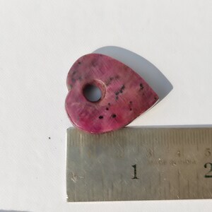 May include: A pink heart-shaped stone with a hole in the center. The stone is about 1 inch wide and is sitting on a ruler.