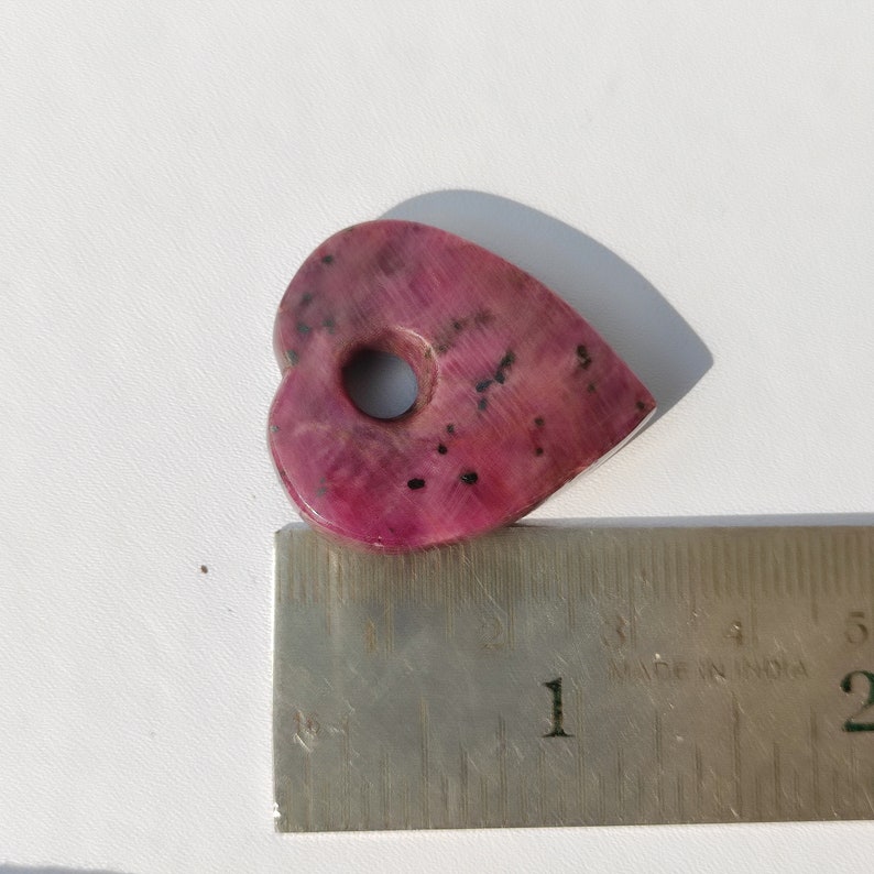 May include: A pink heart-shaped stone with a hole in the center. The stone is about 1 inch wide and is sitting on a ruler.