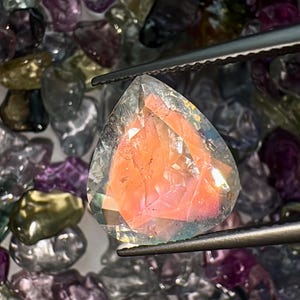 May include: A faceted, clear gemstone with a pink and orange interior. The gemstone is held by tweezers and surrounded by other loose gemstones.