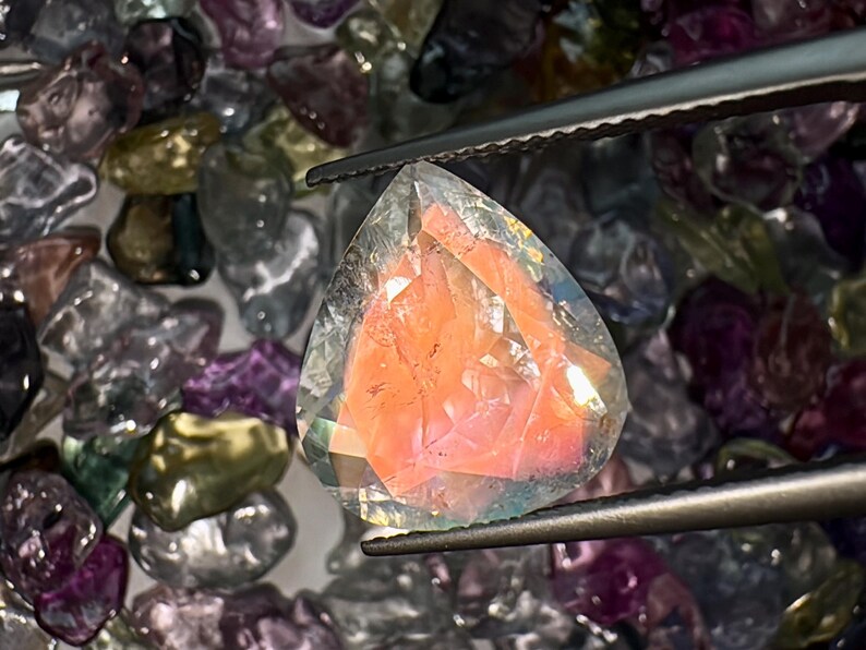 May include: A faceted, clear gemstone with a pink and orange interior. The gemstone is held by tweezers and surrounded by other loose gemstones.