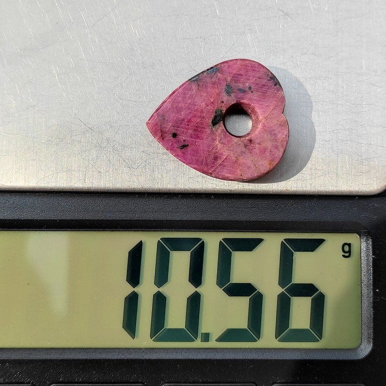 May include: A pink heart-shaped gemstone with a hole in the center, weighing 1.56 grams, on a digital scale.