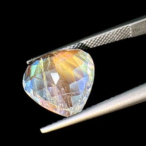 May include: A heart-shaped iridescent gemstone with a rainbow of colors, held by tweezers against a black background.