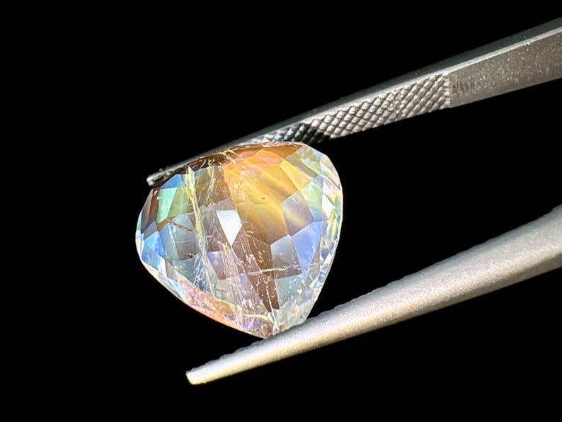 May include: A heart-shaped iridescent gemstone with a rainbow of colors, held by tweezers against a black background.