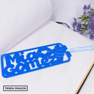 May include: A bright blue glittery bookmark featuring the name Mia Gomez in a cutout font style. The design includes a small ice cream cone icon and a pair of lips. A light blue ribbon is attached to the top right corner of the rectangular bookmark.