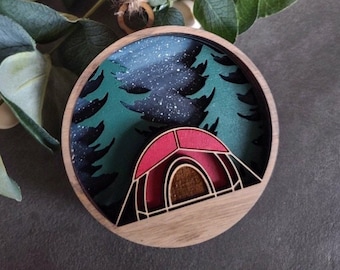 May include: A circular wooden ornament featuring a layered design of a red tent set against a dark green forest background with white speckles representing stars. The piece is framed in a light wood border and includes a wooden bead hanging loop.