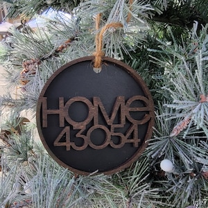May include: A circular wooden ornament featuring a dark brown frame and a black center. The word HOME is cut out in dark brown wood above the numbers 43054. A piece of natural jute twine is looped through a hole at the top for hanging purposes.