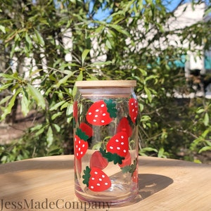 K&ouml;nnte beinhalten: Ein klares Glas mit Bambusdeckel, verziert mit roten Erdbeeren und gr&uuml;nen Bl&auml;ttern. Der Becher ist zylindrisch und steht auf einer Holzoberfl&auml;che. Der Text "JessMadeCompany" ist unten sichtbar.