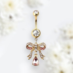 May include: Gold belly button ring with a dangling crystal bow and a teardrop-shaped crystal. The bow is decorated with small pink crystals.