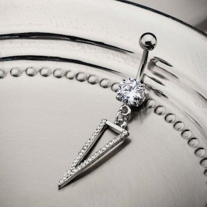 May include: Silver belly button ring with a clear crystal and a dangling triangle charm. The triangle is outlined with smaller crystals.