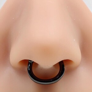 Könnte beinhalten: Ein glatter, kreisförmiger schwarzer Septumring, der in der Nasenscheidewand getragen wird. Das Schmuckstück hat ein durchgehendes, nahtloses Reifendesign mit einer polierten, glänzenden Oberfläche, die sich von der hautfarbenen Nasenattrappe abhebt.