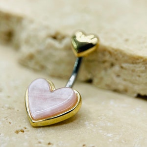 Puede incluir: Un anillo de ombligo de oro con dos dijes en forma de corazón rosa. El dije más grande tiene un borde dorado.