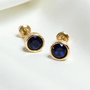 May include: A pair of gold-colored stud earrings with round, bezel-set, deep blue gemstones. The earrings are displayed on a white, textured surface, creating a contrast between the jewelry and the background.