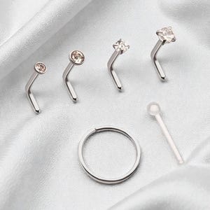Puede incluir: Una variedad de joyas para la nariz, que incluyen cuatro piercings en forma de L con piedras preciosas transparentes y color champán, un aro de nariz plateado y un hueso de nariz de plástico transparente. Presentado sobre tela blanca y sedosa.