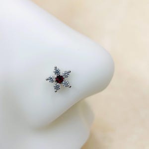 May include: A silver nose ring with a red gemstone in the center and clear rhinestones around it, shaped like a flower.