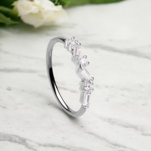 Peut inclure: Une bague en argent au design délicat et incurvé. Elle est ornée d'une série de petites pierres précieuses claires, dont des tailles baguette et rondes, serties sur le dessus. La bague est présentée sur une surface en marbre.