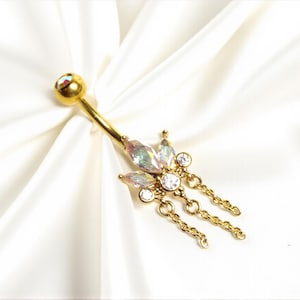 May include: Gold-colored belly button ring with a curved barbell design. The jewelry features iridescent, teardrop-shaped gems and small clear crystals, with dangling chains. The ring is set against a white, silky background.
