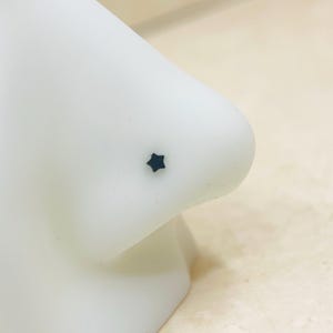 May include: A close-up of a white nose with a small, dark blue star-shaped nose stud. The nose is smooth and appears to be made of a matte material. The background is a light beige color.
