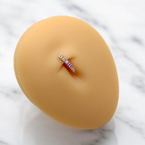 Op de afbeelding: Een navelpiercing met een goudkleurige band en een rij kleurrijke, rechthoekige edelstenen. De ring wordt getoond op een lichtbruine, ovale vorm, tegen een witte en grijze marmeren achtergrond. De edelstenen zijn in roze, paarse en blauwe tinten.