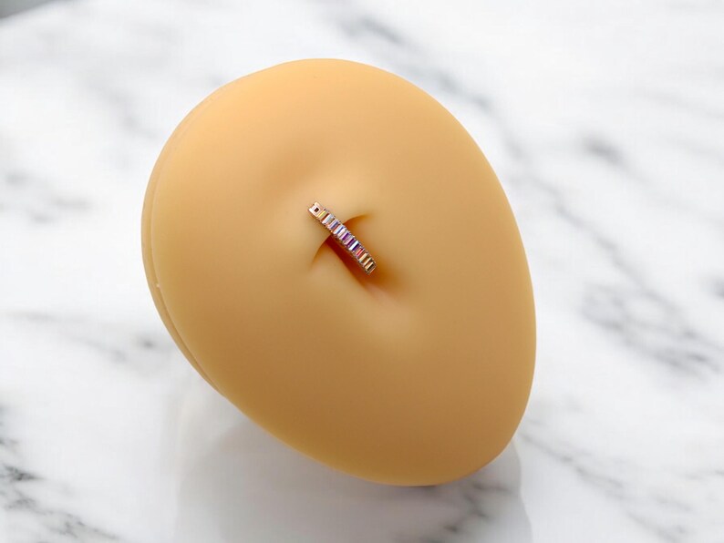 Op de afbeelding: Een navelpiercing met een goudkleurige band en een rij kleurrijke, rechthoekige edelstenen. De ring wordt getoond op een lichtbruine, ovale vorm, tegen een witte en grijze marmeren achtergrond. De edelstenen zijn in roze, paarse en blauwe tinten.