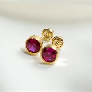 Peut inclure: Une paire de boucles d'oreilles à clous en or avec des pierres précieuses rondes rouge rubis. Les boucles d'oreilles sont serties dans un sertissage simple, mettant en valeur la couleur des pierres. Sur une surface blanche.