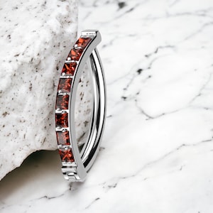 May include: A silver hoop earring with a row of red cubic zirconia stones set in a curved design.