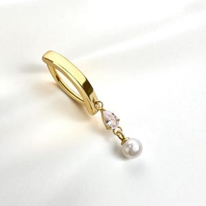 Puede incluir: Un piercing para el ombligo de color dorado con una gema transparente en forma de pera y un pequeño colgante de perla blanca. El anillo está sobre un fondo blanco.