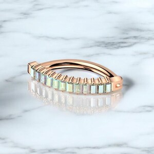 Op de afbeelding: Ros&eacute;gouden ring met een rij rechthoekige, lichtgekleurde edelstenen. De gebogen ring ligt op een wit marmeren oppervlak, met een weerspiegeling van het ontwerp.