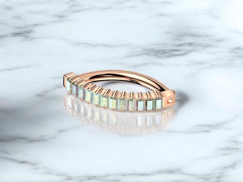 Op de afbeelding: Ros&eacute;gouden ring met een rij rechthoekige, lichtgekleurde edelstenen. De gebogen ring ligt op een wit marmeren oppervlak, met een weerspiegeling van het ontwerp.
