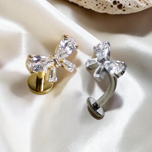 May include: Two bow-shaped body jewelry pieces featuring clear, faceted stones. One piece has a gold-toned flat back post, while the other has a silver-toned curved post. Both items are designed with a central bow motif adorned with sparkling crystals.