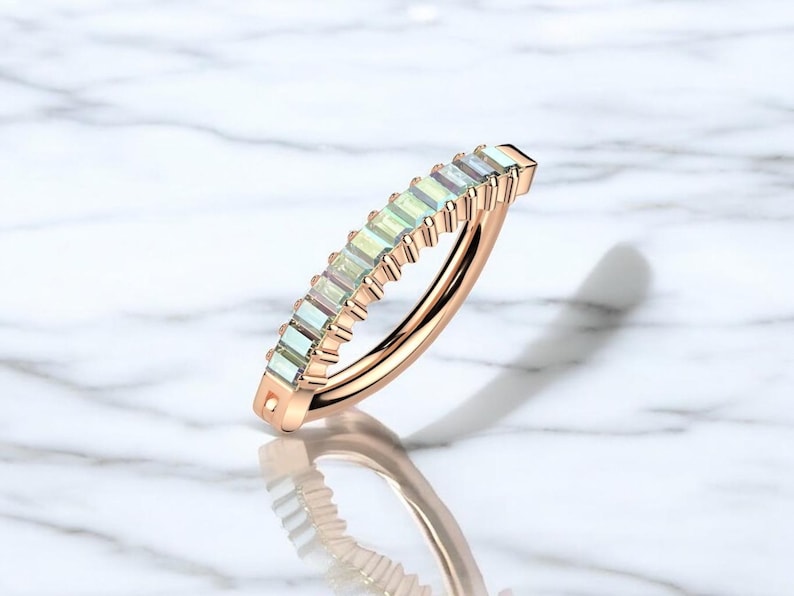 Op de afbeelding: Ros&eacute;gouden ring met een rij rechthoekige, lichtgroene edelstenen. De ring is geplaatst tegen een witte marmeren achtergrond, met een schaduw eronder. De edelstenen zijn gelijkmatig verdeeld.