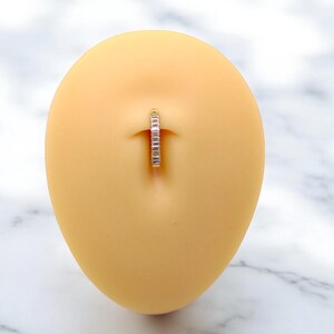 Op de afbeelding: Een navelpiercing met een zilverkleurige ring en kleine, heldere steentjes. De sieraden worden gepresenteerd op een lichtgele, ovaalvormige vorm, tegen een witte en grijze marmeren achtergrond. De ring is ontworpen voor een navelpiercing.