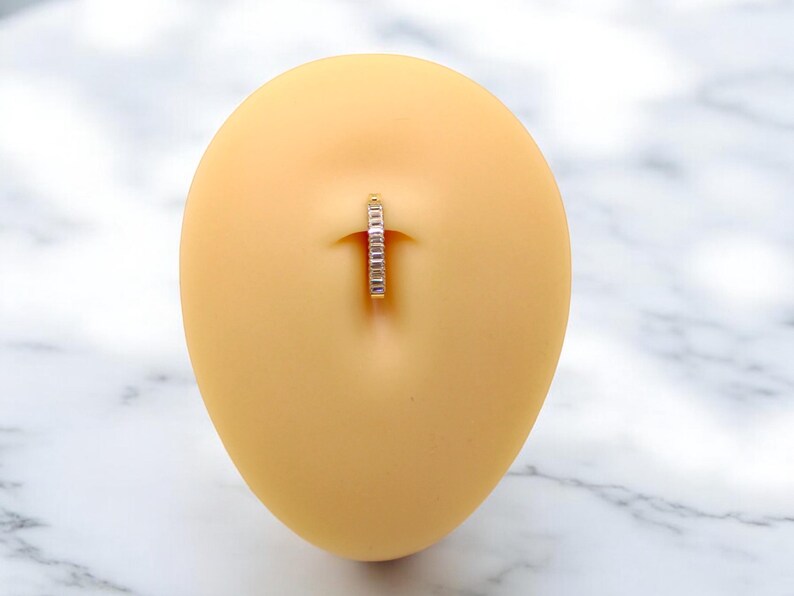 Op de afbeelding: Een navelpiercing met een zilverkleurige ring en kleine, heldere steentjes. De sieraden worden gepresenteerd op een lichtgele, ovaalvormige vorm, tegen een witte en grijze marmeren achtergrond. De ring is ontworpen voor een navelpiercing.