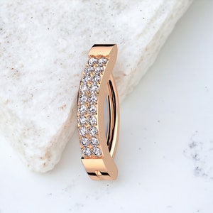 May include: A rose gold body jewellery ring with a curved shape and two rows of clear cubic zirconia stones.
