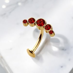 May include: Gold body jewellery with a curved bar and five red gemstones. The gemstones are arranged in a crescent shape.