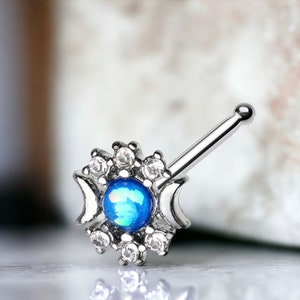 Puede incluir: Un aro de nariz de plata con una piedra de ópalo azul rodeada de piedras de circonita cúbica transparentes. El aro tiene un diseño de media luna.