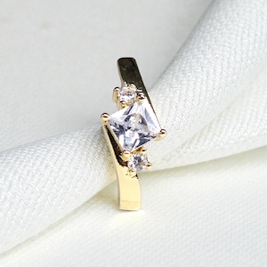May include: A gold-colored earring with a square, clear gemstone at the center, flanked by two smaller, round, clear gemstones. The earring is set against a white, textured background.