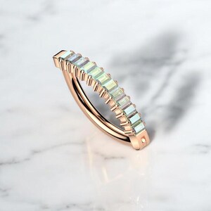 Op de afbeelding: Ros&eacute;gouden ring met een rij rechthoekige, iriserende edelstenen. De band van de ring buigt sierlijk, waarbij de kleurvariaties van de edelstenen worden getoond. De ring is geplaatst op een wit marmeren oppervlak.