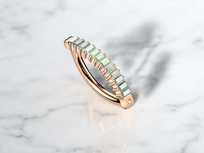 Op de afbeelding: Ros&eacute;gouden ring met een rij rechthoekige, iriserende edelstenen. De band van de ring buigt sierlijk, waarbij de kleurvariaties van de edelstenen worden getoond. De ring is geplaatst op een wit marmeren oppervlak.