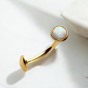 May include: Gold body jewelry with a white opal gemstone. The jewelry is a curved barbell style.