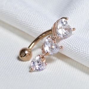 May include: A rose gold belly button ring with three clear, heart-shaped cubic zirconia stones. The curved barbell design features a small ball at the bottom and a larger heart-shaped stone at the top, with two more hearts dangling below.