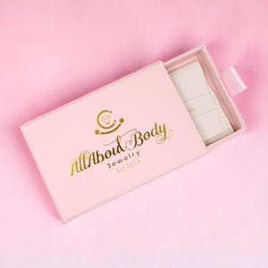 Op de afbeelding: Een lichtroze sieradendoosje met de gouden tekst "All About Body Jewelry Est. 2019". De doos is gedeeltelijk open en toont een wit interieur en een wit lint. De achtergrond is effen roze.