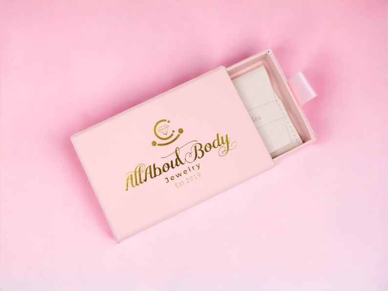 Op de afbeelding: Een lichtroze sieradendoosje met de gouden tekst "All About Body Jewelry Est. 2019". De doos is gedeeltelijk open en toont een wit interieur en een wit lint. De achtergrond is effen roze.