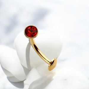 Pode incluir: Joia de corpo dourada com uma pedra preciosa vermelha. A joia é curva e tem um verso plano.